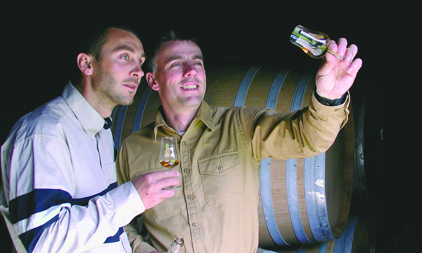 Joël et Olivier Billy examinant une eau-de-vie devant un fût, dans le chai de la propriété
